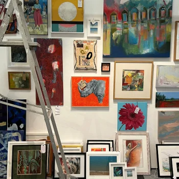 Wall in an art gallery densely hung with assorted framed artworks in bright colours; a metal ladder leans on the left.