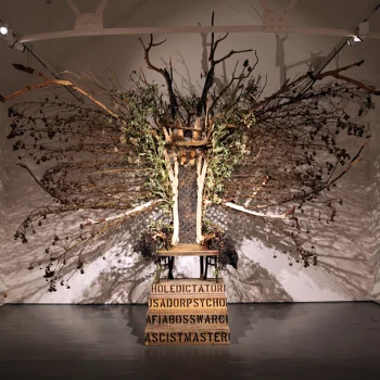Large bare-tree sculpture in a gallery, branches radiating from a central trunk over a wooden platform, atop a crate with bold lettering.