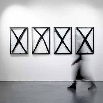 Four framed artworks with bold black Xs on white panels hung on a gallery wall; a blurred figure walks past.