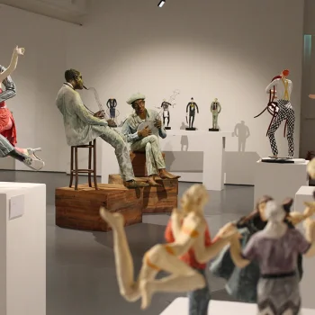 Gallery of life-size sculptures: two seated figures on wooden blocks, one with a hat playing a guitar; several statues on pedestals, foreground figures blurred.