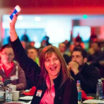 A person in the foreground raises a hand holding a phone, smiling; others clap at tables with bottles under red and blue lighting in a crowded event.