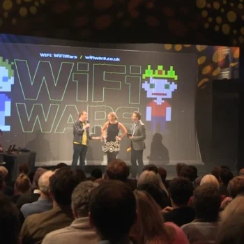 Three presenters on a stage in front of a large WiFi Wars screen with pixel-art figures; audience watches as side screens show circular graphics.