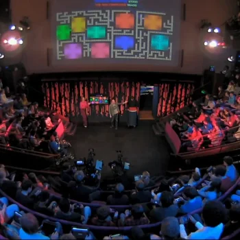 Wide shot of a TV studio with a stage; three people on stage as a large screen shows a colourful Pac-Man maze, with audience in tiered seating.