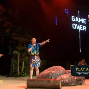 Person in a tropical shirt and shorts on stage with a microphone, pointing; neon "GAME OVER" scoreboard behind, gold pillars and large props, "PLAY ALONG" banner.
