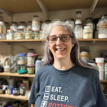 Smiling person with grey hair and red-framed glasses, wearing a dark T-shirt reading “EAT. SLEEP. POTTERY. REPEAT.”, in a pottery studio with shelves of glaze jars.