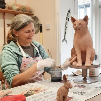 Smiling potter in a patterned apron and gloves shapes a large terracotta cat on a raised stand, with a small rabbit sculpture on a newspaper-covered table.