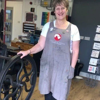 Person with short hair wearing a paint-splattered grey apron over a white top stands in a workshop, resting a hand on a large black metal wheel.