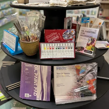 Round black display stand in a shop filled with art supplies: brushes in a pot, watercolor sets, a Seawhite paint box, and purple pads.