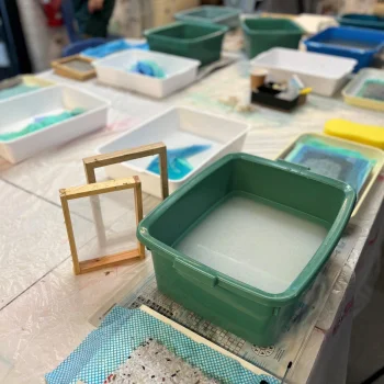 Craft workshop table with a large green tub of water, white and coloured trays, wooden frames, a blue speckled sheet, and blurred figures in aprons.