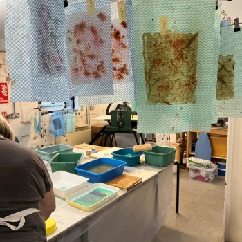 Art studio with blue chevron sheets clipped to a line, stained with dye. A long table covered in plastic holds water trays and bins; a person in an apron stands at the left.