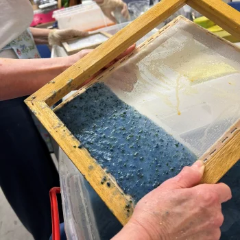 Hands hold a wooden-framed silkscreen with mesh; blue speckled pigment covers the lower half, with a pale yellow area on the upper section, in a workshop.