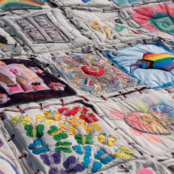 Patchwork quilt on pale fabric with embroidered squares showing butterflies, flowers, rainbows, mandalas and figures, tied with red threads.