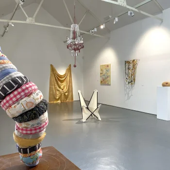 In a white-walled gallery, a tall stack of fabric rolls on the left, a gold draped sculpture, a pink chandelier, and a pedestal with a doughnut-like object.