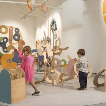 Two children explore a large abstract wooden sculpture made of colourful curved shapes in a bright gallery.