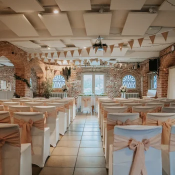 Brick-walled wedding venue with white chairs, peach sashes, bunting, string lights and a central aisle to floral arrangements by arched windows.
