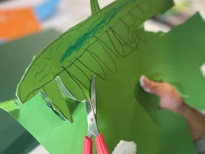 An image featuring a child's hands, they are cutting a green piece of card with scissors in their left hand. The card has a sea creature drawn on.