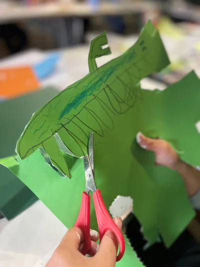 An image featuring a child's hands, they are cutting a green piece of card with scissors in their left hand. The card has a sea creature drawn on.
