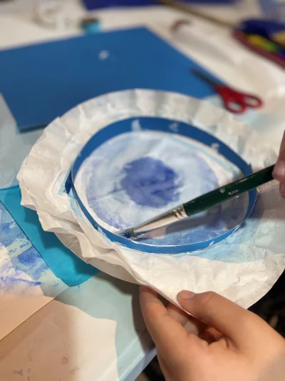 An image of a child's hands, they are painting a piece of circular tissue paper blue to become a sea creature of their design.