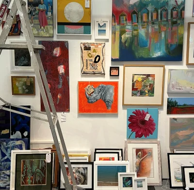 Wall in an art gallery densely hung with assorted framed artworks in bright colours; a metal ladder leans on the left.