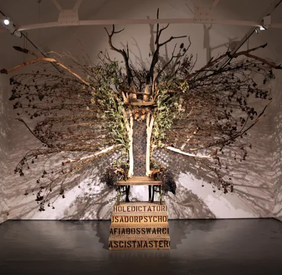 Large bare-tree sculpture in a gallery, branches radiating from a central trunk over a wooden platform, atop a crate with bold lettering.