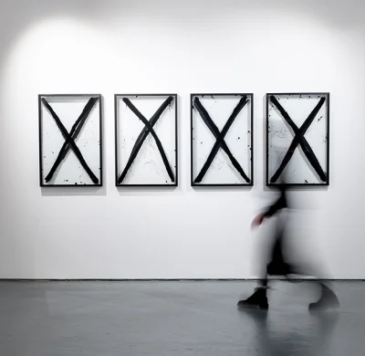 Four framed artworks with bold black Xs on white panels hung on a gallery wall; a blurred figure walks past.
