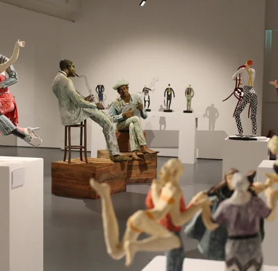 Gallery of life-size sculptures: two seated figures on wooden blocks, one with a hat playing a guitar; several statues on pedestals, foreground figures blurred.