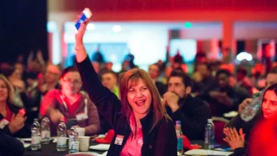 A person in the foreground raises a hand holding a phone, smiling; others clap at tables with bottles under red and blue lighting in a crowded event.
