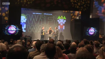 Three presenters on a stage in front of a large WiFi Wars screen with pixel-art figures; audience watches as side screens show circular graphics.