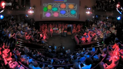 Wide shot of a TV studio with a stage; three people on stage as a large screen shows a colourful Pac-Man maze, with audience in tiered seating.