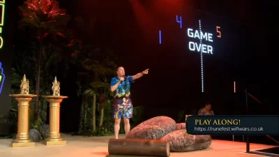 Person in a tropical shirt and shorts on stage with a microphone, pointing; neon "GAME OVER" scoreboard behind, gold pillars and large props, "PLAY ALONG" banner.