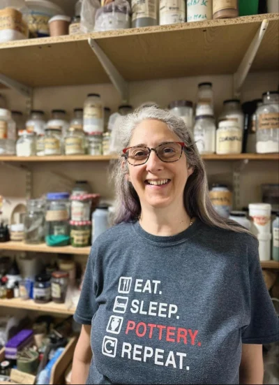 Smiling person with grey hair and red-framed glasses, wearing a dark T-shirt reading “EAT. SLEEP. POTTERY. REPEAT.”, in a pottery studio with shelves of glaze jars.