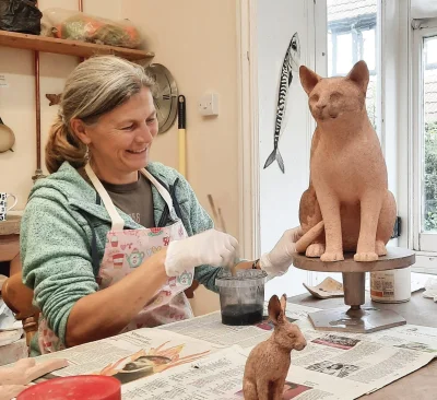Smiling potter in a patterned apron and gloves shapes a large terracotta cat on a raised stand, with a small rabbit sculpture on a newspaper-covered table.