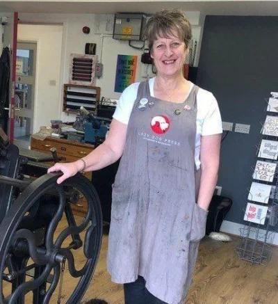 Person with short hair wearing a paint-splattered grey apron over a white top stands in a workshop, resting a hand on a large black metal wheel.