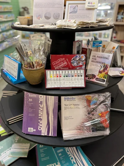 Round black display stand in a shop filled with art supplies: brushes in a pot, watercolor sets, a Seawhite paint box, and purple pads.