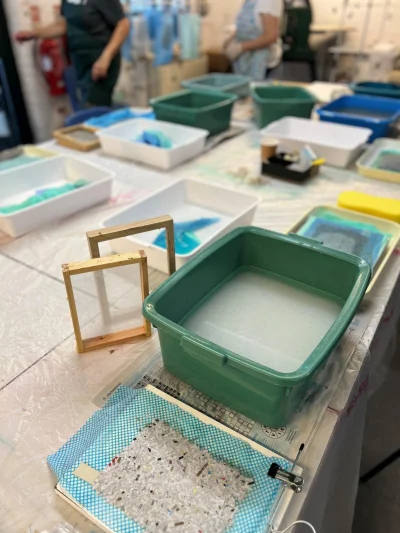 Craft workshop table with a large green tub of water, white and coloured trays, wooden frames, a blue speckled sheet, and blurred figures in aprons.