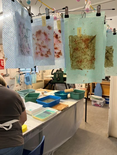 Art studio with blue chevron sheets clipped to a line, stained with dye. A long table covered in plastic holds water trays and bins; a person in an apron stands at the left.