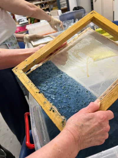 Hands hold a wooden-framed silkscreen with mesh; blue speckled pigment covers the lower half, with a pale yellow area on the upper section, in a workshop.