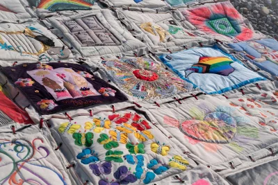 Patchwork quilt on pale fabric with embroidered squares showing butterflies, flowers, rainbows, mandalas and figures, tied with red threads.