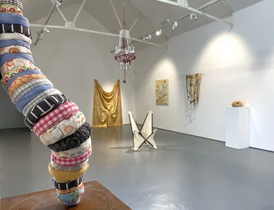 In a white-walled gallery, a tall stack of fabric rolls on the left, a gold draped sculpture, a pink chandelier, and a pedestal with a doughnut-like object.