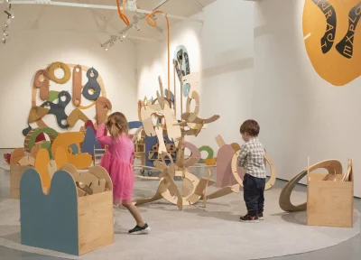 Two children explore a large abstract wooden sculpture made of colourful curved shapes in a bright gallery.