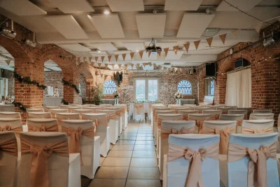 Brick-walled wedding venue with white chairs, peach sashes, bunting, string lights and a central aisle to floral arrangements by arched windows.