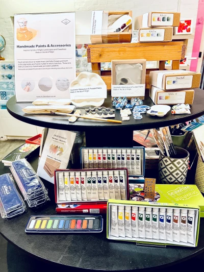 Round black display table filled with art supplies: watercolor tubes, palettes, brushes, boxed paints, and a sign reading Handmade Paints & Accessories.