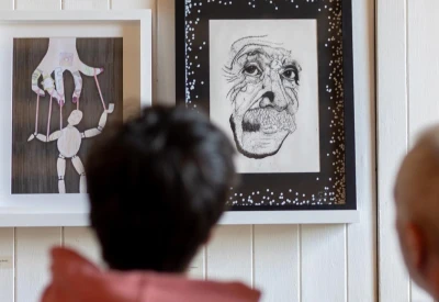 Two framed artworks on a white wall; left shows a puppet figure, right a detailed elderly face with a speckled border; blurred heads in foreground.