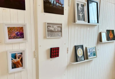 White paneled gallery wall with multiple framed artworks on the wall and shelves, including colorful floral photos and a grayscale car image.