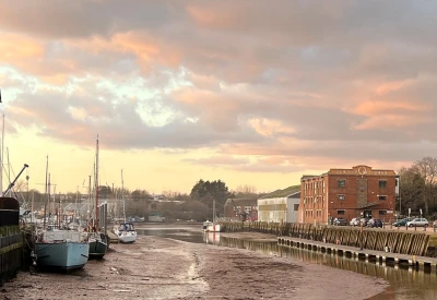 Sunset over a harbor with moored boats on the left, a brick Jubilee Stores building on the right, and a calm waterway reflecting a pink-orange sky.