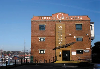 Brick building by a marina with a vertical STUDIOS banner and a Jubilee Stores sign on the roof; boats and water to the left.