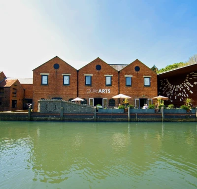 Brick building with three gables labelled QUAY ARTS beside canal, outdoor seating under umbrellas on terrace, and a wooden wall with art to the right.