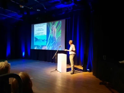 Speaker stands at a white podium on a dim stage with blue lighting; a projected slide with text and images is displayed on the screen beside them.
