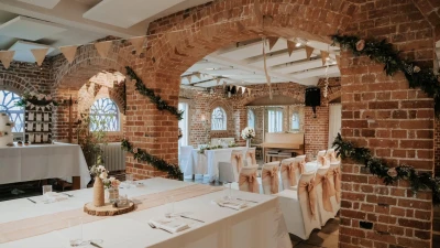 Rustic wedding reception in a brick-arched hall with white-clothed tables, chairs in peach sashes, floral garlands, bunting and warm fairy lights.