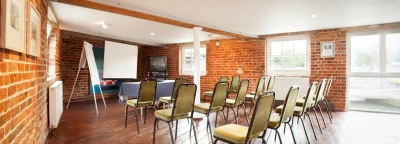 Brick-walled meeting room with a wooden floor, rows of beige chairs facing a projector screen and whiteboard, a table at the front and large windows.